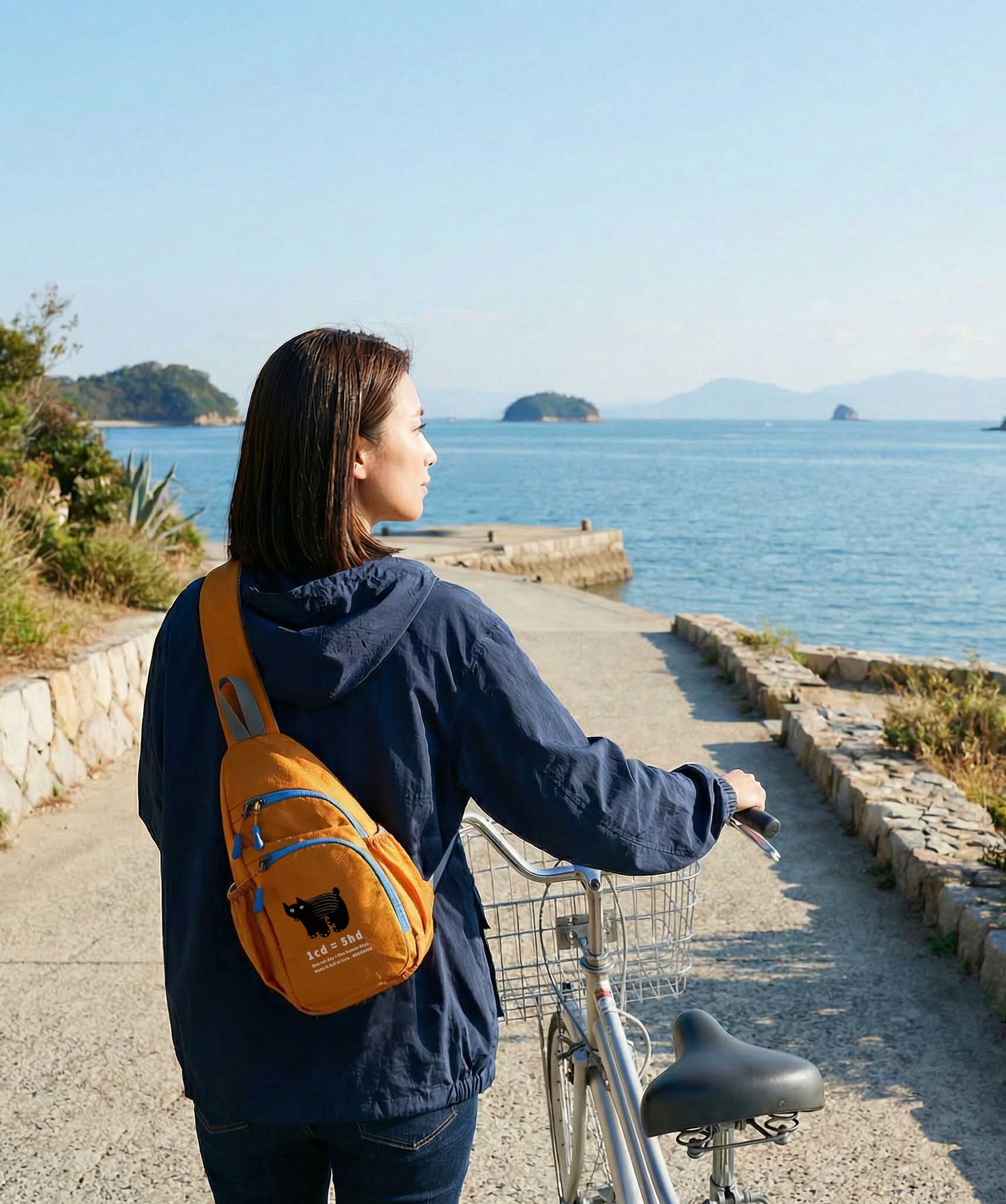 瀬戸内の海沿いを自転車で走る女性・オレンジのNEKONI SLINGバッグ