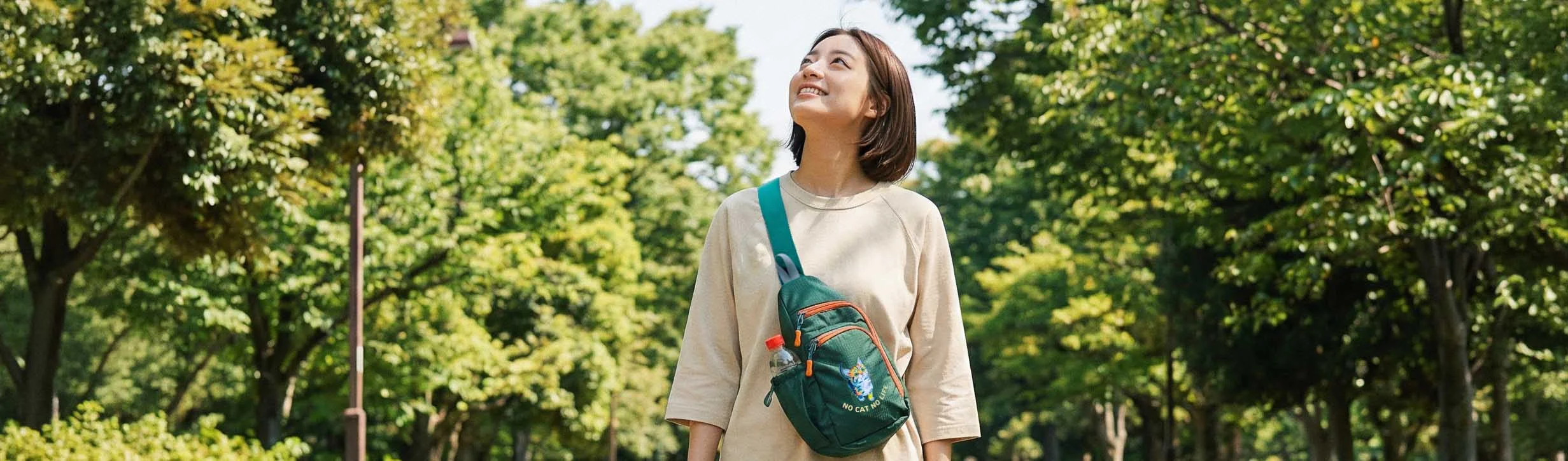 公園を歩く女性・グリーンのNEKONI SLINGバッグ・空を見上げる